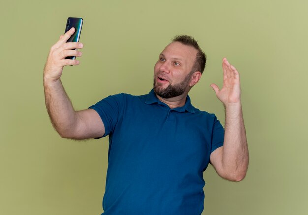 Impressed adult slavic man holding and looking at mobile phone and keeping hand in air 