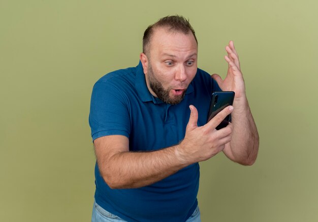 Impressed adult slavic man holding and looking at mobile phone keeping hand in air 