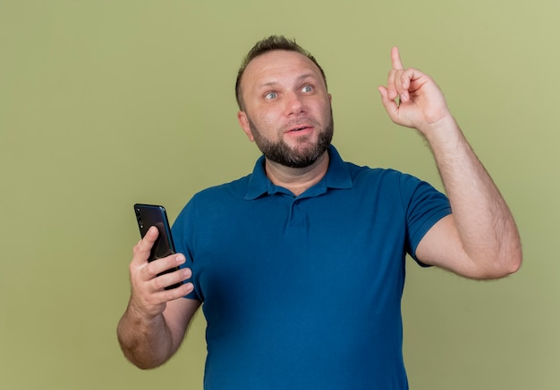 Впечатленный взрослый славянский мужчина держит мобильный телефон, глядя в сторону и поднимая палец