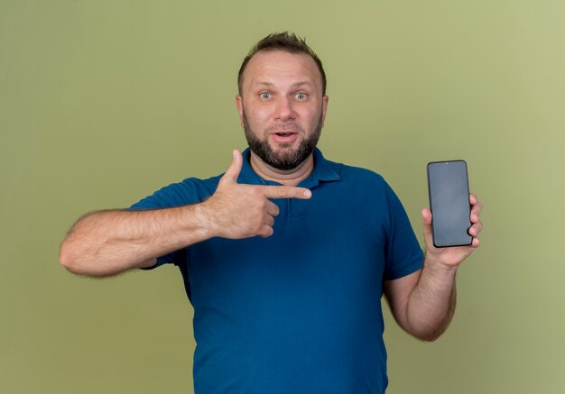 Impressed adult slavic man holding and pointing at mobile phone looking 