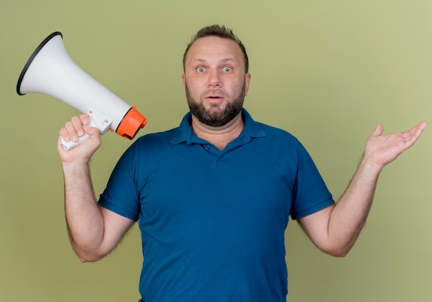 Impressed adult slavic man holding speaker and showing empty hand 