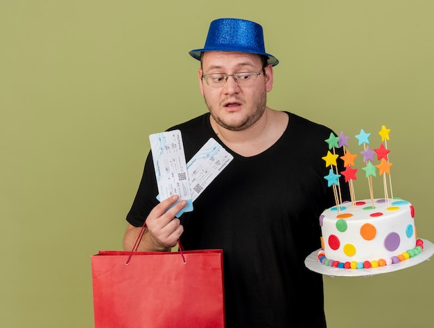 Бесплатное фото Впечатленный взрослый славянский мужчина в оптических очках в синей шляпе держит бумажную сумку, торт ко дню рождения и авиабилеты