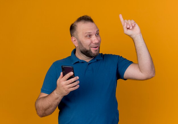 Impressed adult slavic man looking at side holding mobile phone raising finger 