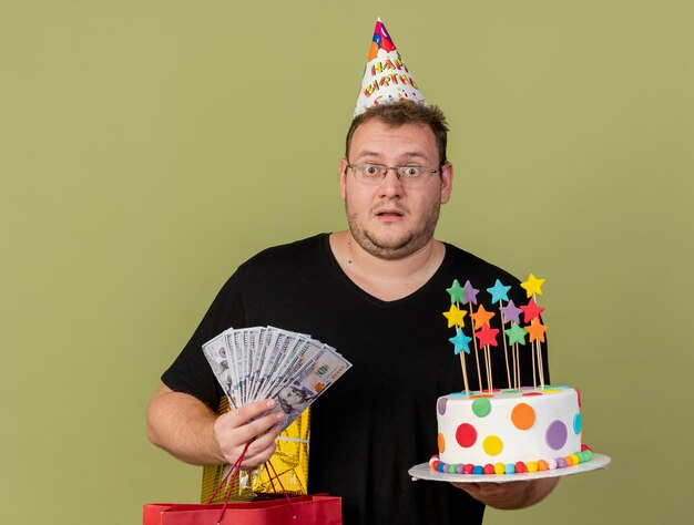 Impressed adult slavic man in optical glasses wearing birthday cap holds money paper shopping bag gift box and birthday cake 