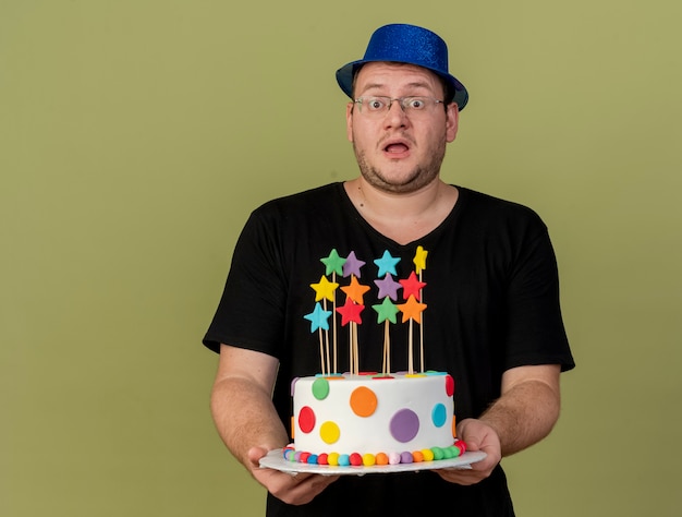 Impressed adult slavic man in optical glasses wearing blue party hat holds birthday cake 