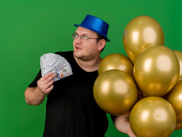 Impressed adult slavic man in optical glasses wearing blue party hat holds helium balloons and money looking at side 