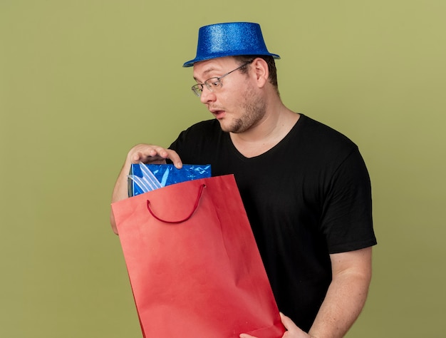 Impressed adult slavic man in optical glasses wearing blue party hat holds and looks at gift box in paper shopping bag 