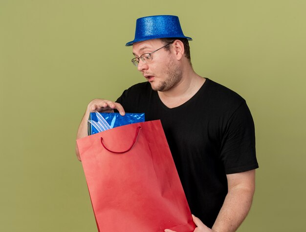 파란색 파티 모자를 쓰고 광학 안경에 인상적인 성인 슬라브 남자가 종이 쇼핑백에 선물 상자를 들고 보인다.