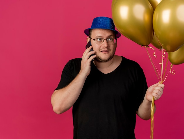 Impressionato uomo slavo adulto in occhiali ottici che indossa un cappello da festa blu tiene e guarda palloncini di elio che parlano al telefono