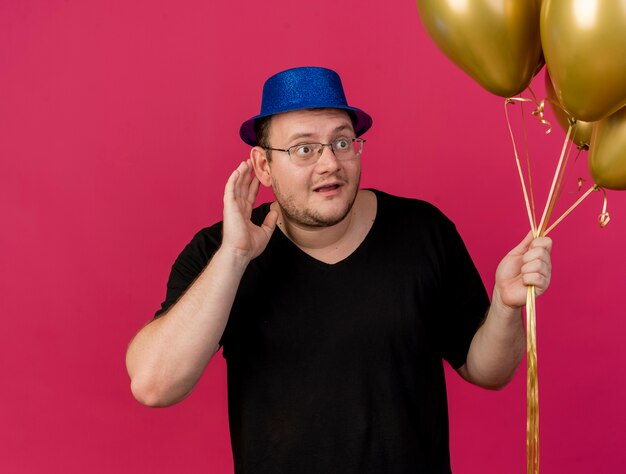 Impressed adult slavic man in optical glasses wearing blue party hat keeps hand close to ear trying to hear and holds helium balloons 