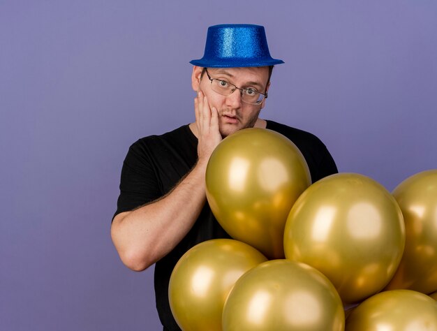 파란색 파티 모자를 쓰고 광학 안경을 쓴 성인 슬라브 남자가 얼굴에 손을 대고 헬륨 풍선으로 서 있습니다.