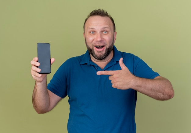 Impressed adult slavic man showing mobile phone and pointing at it 