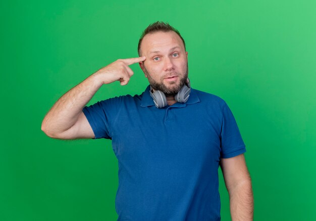 Impressed adult slavic man wearing headphones on neck looking and pointing finger on head 