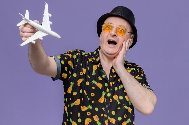 Impressed adult slavic man with black top hat wearing sunglasses putting hand on his face and holding plane model