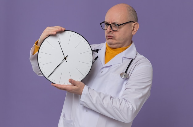 Впечатленный взрослый славянский мужчина в оптических очках в униформе врача со стетоскопом, держащим и смотрящим на часы