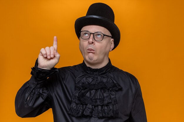 Впечатленный взрослый славянский мужчина в цилиндре и оптических очках в черной готической рубашке смотрит и указывает вверх