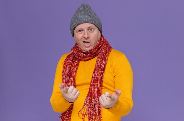 Impressed adult slavic man with winter hat and scarf around his neck looking and pointing at camera 