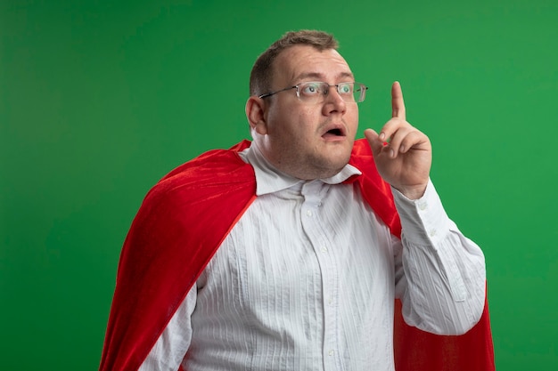 Бесплатное фото Впечатленный взрослый славянский супергерой в красном плаще в очках, глядя прямо, поднимая палец, изолированный на зеленой стене