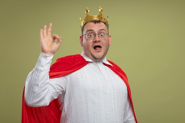 Free photo impressed adult slavic superhero man in red cape wearing glasses and crown  doing ok sign isolated on olive green wall with copy space