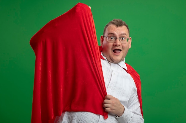 Free photo impressed adult slavic superhero man in red cape wearing glasses grabbing cape looking at camera isolated on green background with copy space