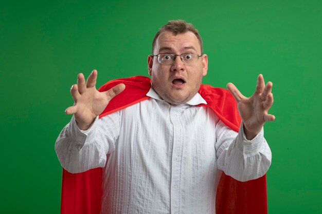 Impressed adult slavic superhero man in red cape wearing glasses  stretching out hands  isolated on green wall