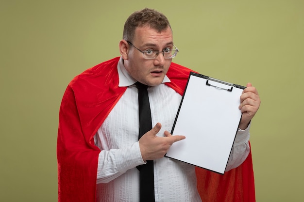 Impressed adult slavic superhero man in red cape wearing glasses and tie holding and pointing at clipboard  isolated on olive green wall