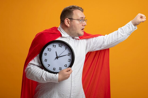 Impressed adult superhero man in red cape wearing glasses holding clock stretching out fist looking at side isolated on orange wall