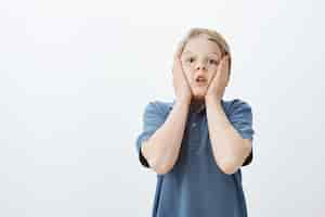 Free photo impressed amazed cute male child in blue t-shirt, holding palms on cheeks, holding breath and gasping from shock and surprise