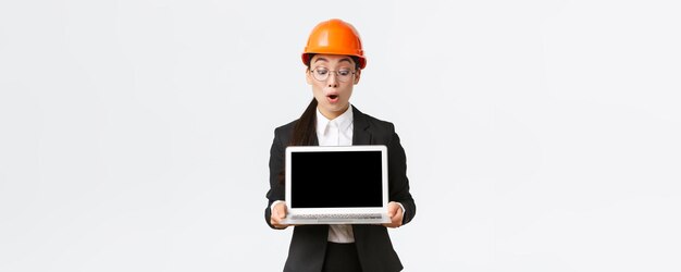 Impressed asian female construction engineer architect or manager at enterprise showing laptop screen with amazed expression looking at profit diagram during meeting wear safety helmet in factory