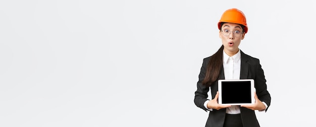 Впечатленная азиатская женщина-строитель, бизнесвумен в защитном шлеме и костюме, показывающая диаграмму, представляет биграмму на экране цифрового планшета, стоя в изумлении на белом фоне