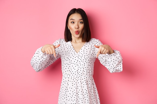 Impressed asian woman in dress pointing fingers down, look there gesture, check out discounts and sales, standing amazed over pink background