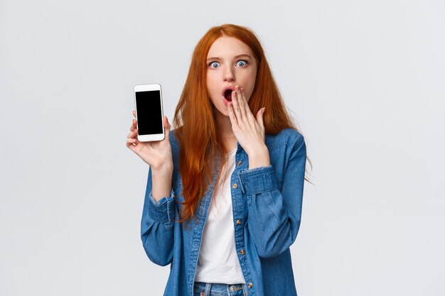 Impressed and astonished redhead woman seeing something unbelievable, spilling tea in internet, gasping cover opened mouth and showing samrtphone display, white background