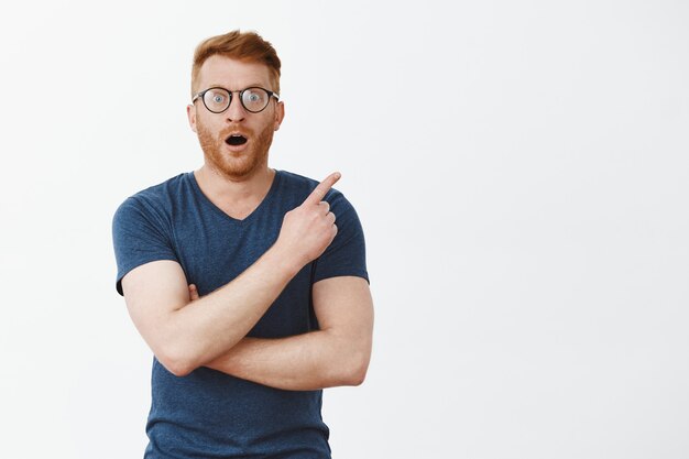 Impressed attractive Caucasian guy with bristle in glasses and blue t-shirt, pointing at upper right corner, gasping and dropping haw from astonishment