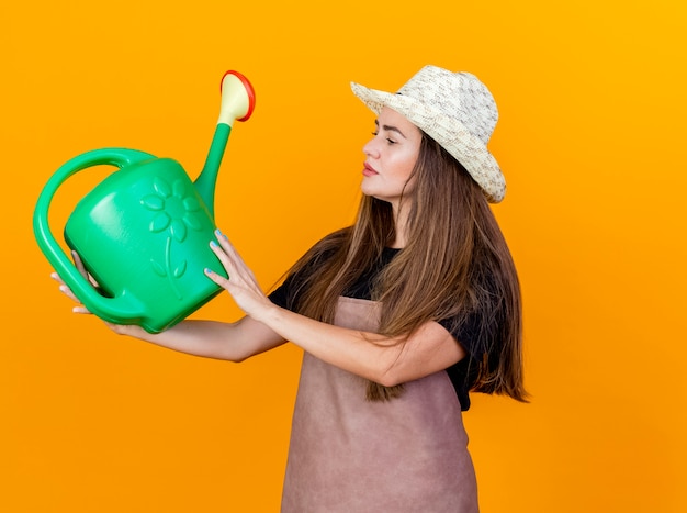 Impressed beautiful gardener girl wearing uniform and gardening hat holding and looking at watering can isolated on orange background