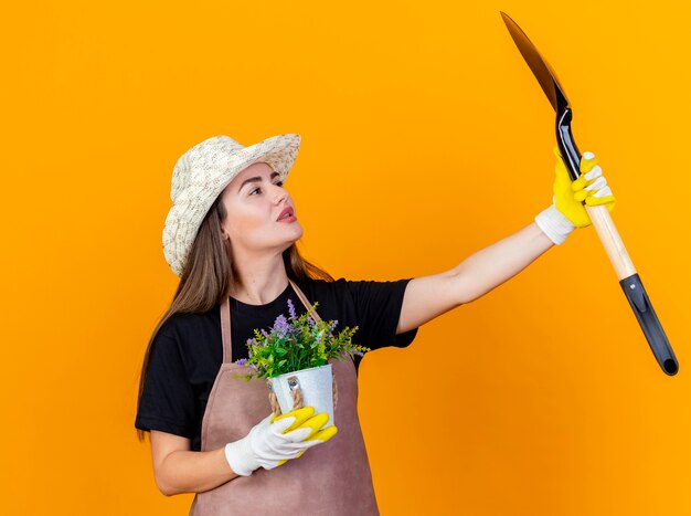 Impressed beautiful gardener girl wearing uniform and gardening hat with gloves holding flower in flowerpot