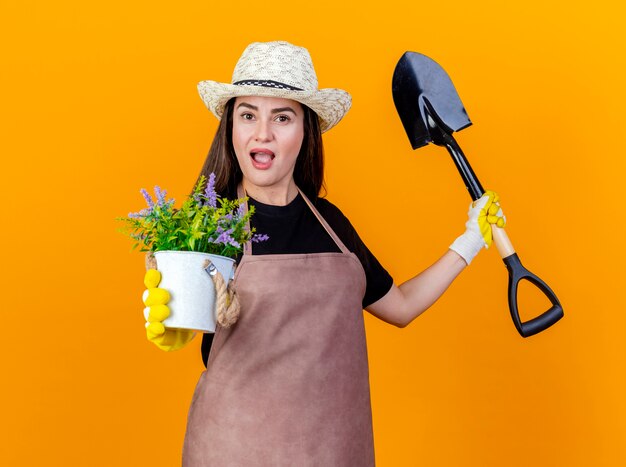 Impressed beautiful gardener girl wearing uniform and gardening hat with gloves holding spade and holding out flower in flowerpot at camera isolated on orange background