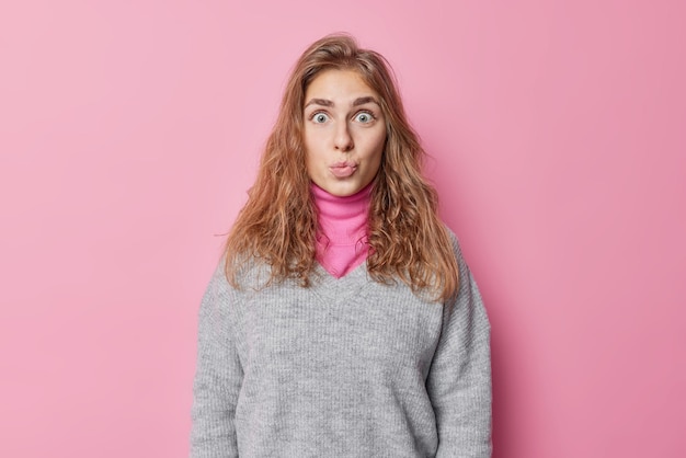 Impressed beautiful long haired young woman keeps lips folded looks with wondered expression has eyes popped out wears warm jumper isolated over pink background awaits for kiss from someone.