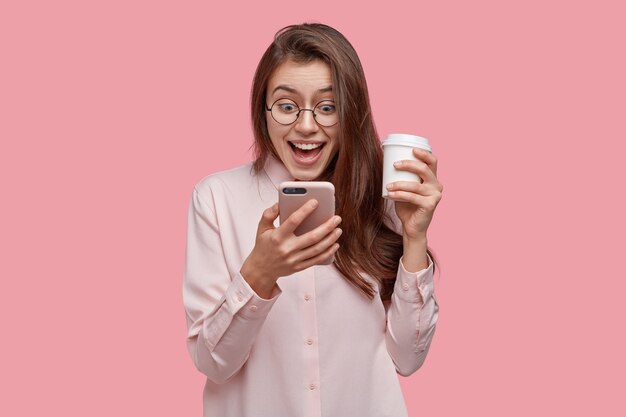 Impressed beautiful woman feels overjoyed, reads text message from excitement, dressed formally, carries takeaway coffee, isolated over pink studio wall. People