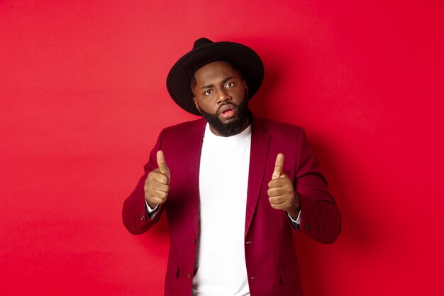 Impressed Black man saying good job, cheer up with thumbs-up, praise something, standing against red background