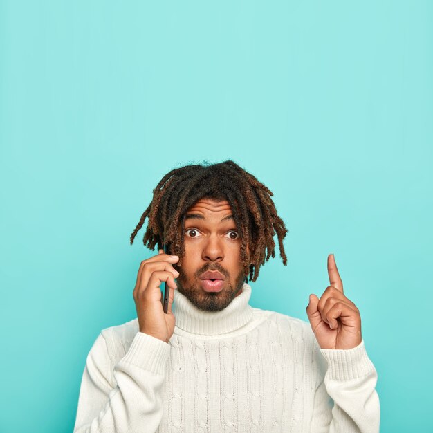 험 상을 가진 인상적인 흑인 남자는 전화 대화, 검지 손가락으로 위의 포인트, 파란색 배경 위에 절연 흰색 캐주얼 스웨터를 입고 텍스트 복사 공간
