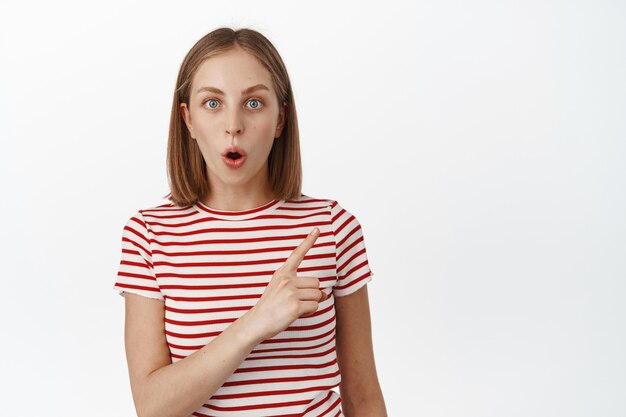 Impressed blond young woman gasp, pointing at upper right corner discount banner, showing company logo, standing in striped summer t-shirt over white wall