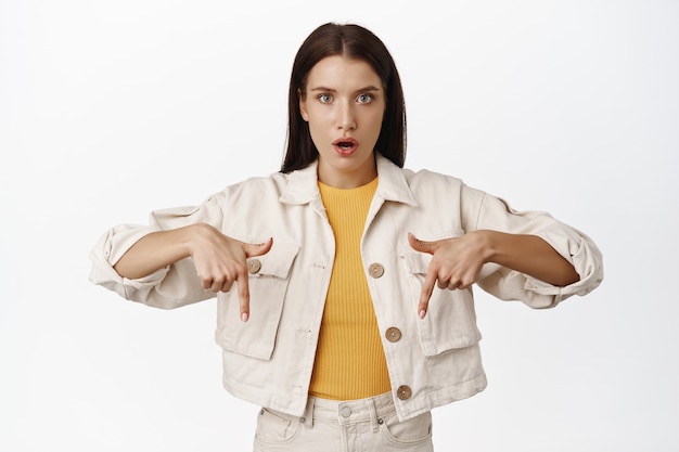 Impressed brunette woman talks about advertisement, pointing fingers down at copy space with sale discount logo, showing banner, open mouth fascinated on white.