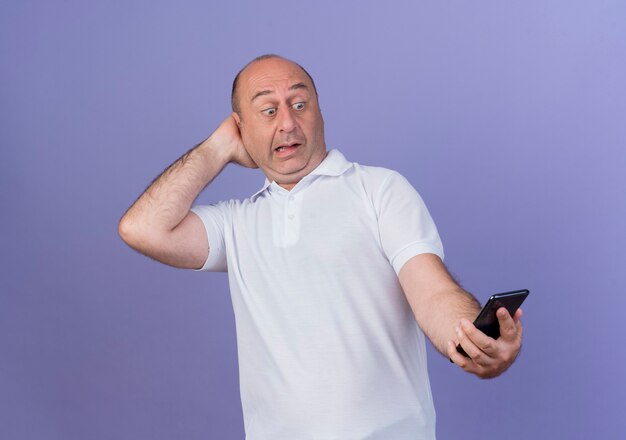 Impressed casual mature businessman holding and looking at mobile phone and putting hand behind head isolated on purple background