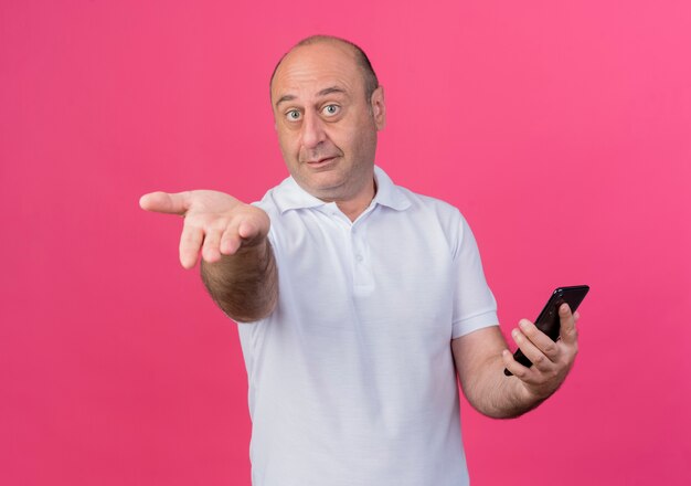 Impressed casual mature businessman holding mobile phone looking at camera and showing empty hand at camera isolated on pink background