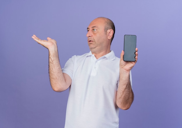 Впечатлен случайный зрелый бизнесмен, держащий мобильный телефон, смотрящий в сторону и показывающий пустую руку, изолированную на фиолетовом фоне с копией пространства
