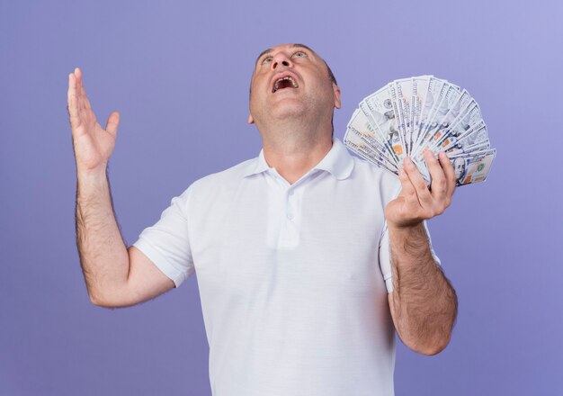 Impressed casual mature businessman holding money keeping hand in air and looking up isolated on purple background