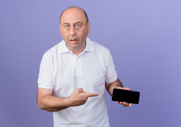 Impressed casual mature businessman holding and pointing at mobile phone isolated on purple background