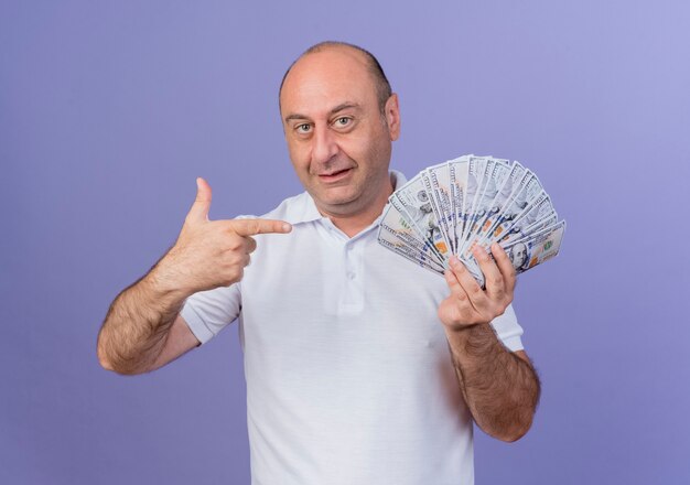 впечатлен случайный зрелый бизнесмен, держащий и указывающий на деньги, изолированные на фиолетовом