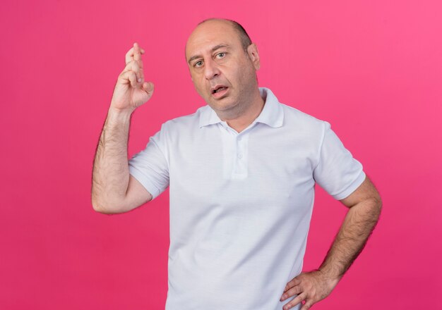 Impressed casual mature businessman keeping hand on waist and crossing fingers isolated on pink background