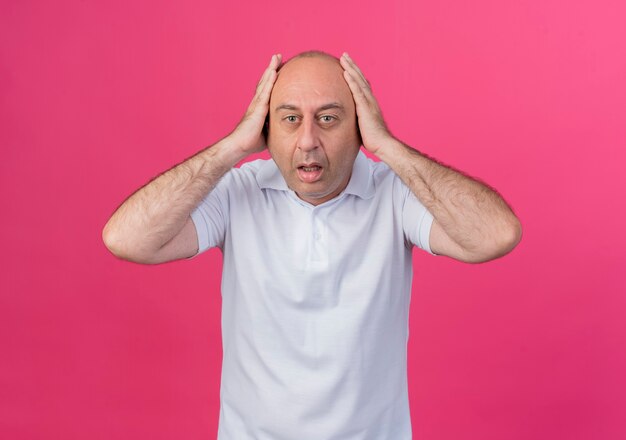 Impressed casual mature businessman looking at camera and holding head isolated on pink background